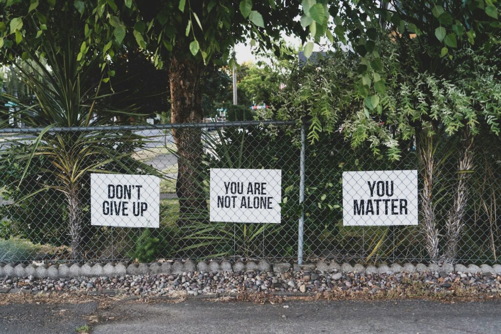 Drei Schilder an einem Zaun mit den Aufschriften 'Don't Give Up', 'You Are Not Alone' und 'You Matter', symbolisierend Unterstützung und Ermutigung.