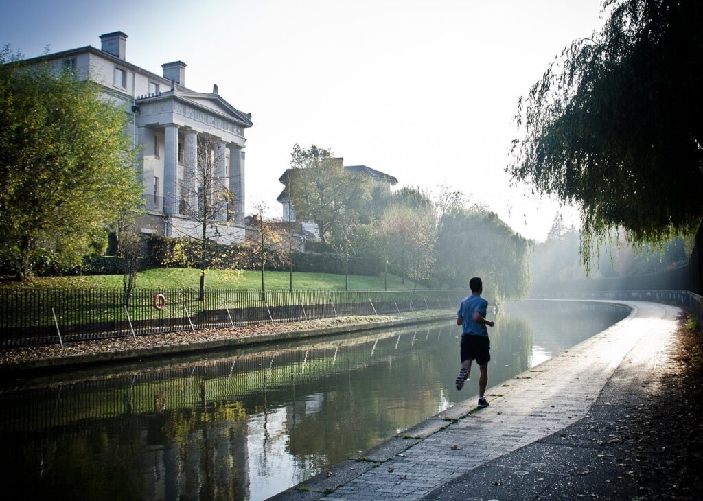 Ein Jogger läuft entlang eines ruhigen Kanals bei Morgenlicht, umgeben von Bäumen und einem historischen Gebäude im Hintergrund.