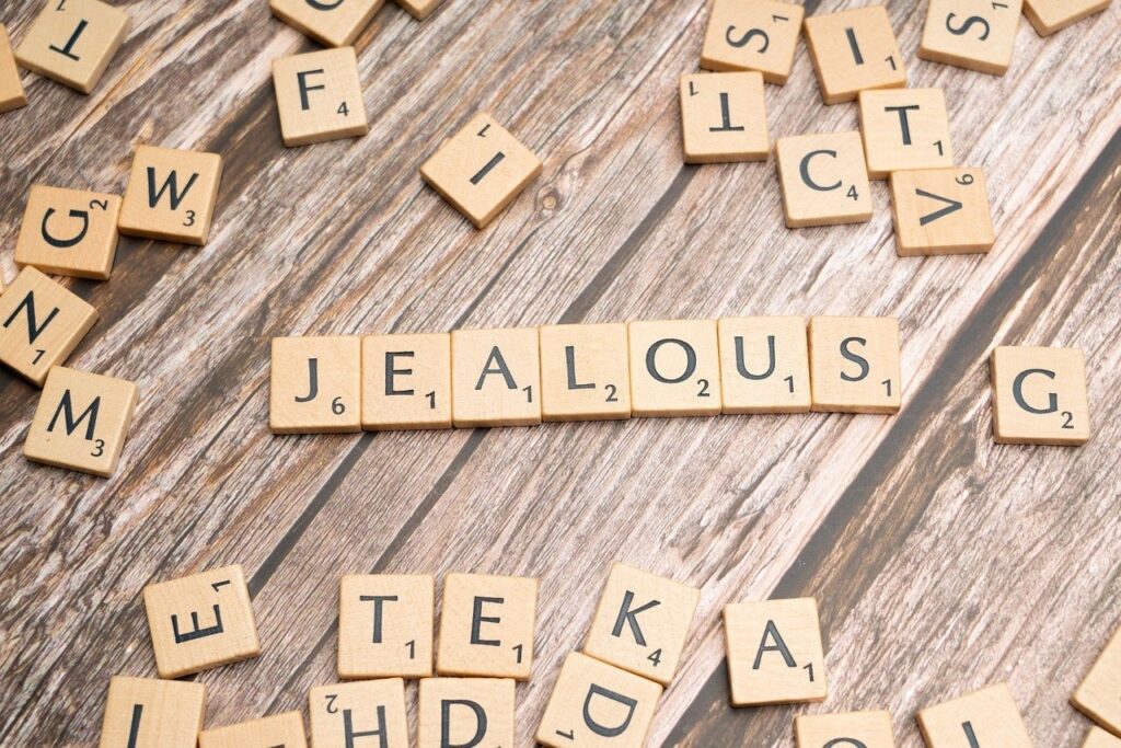 Scrabble-Buchstaben auf einer Holzoberfläche, das Wort 'Jealous' steht im Mittelpunkt, umgeben von verstreuten Buchstaben.